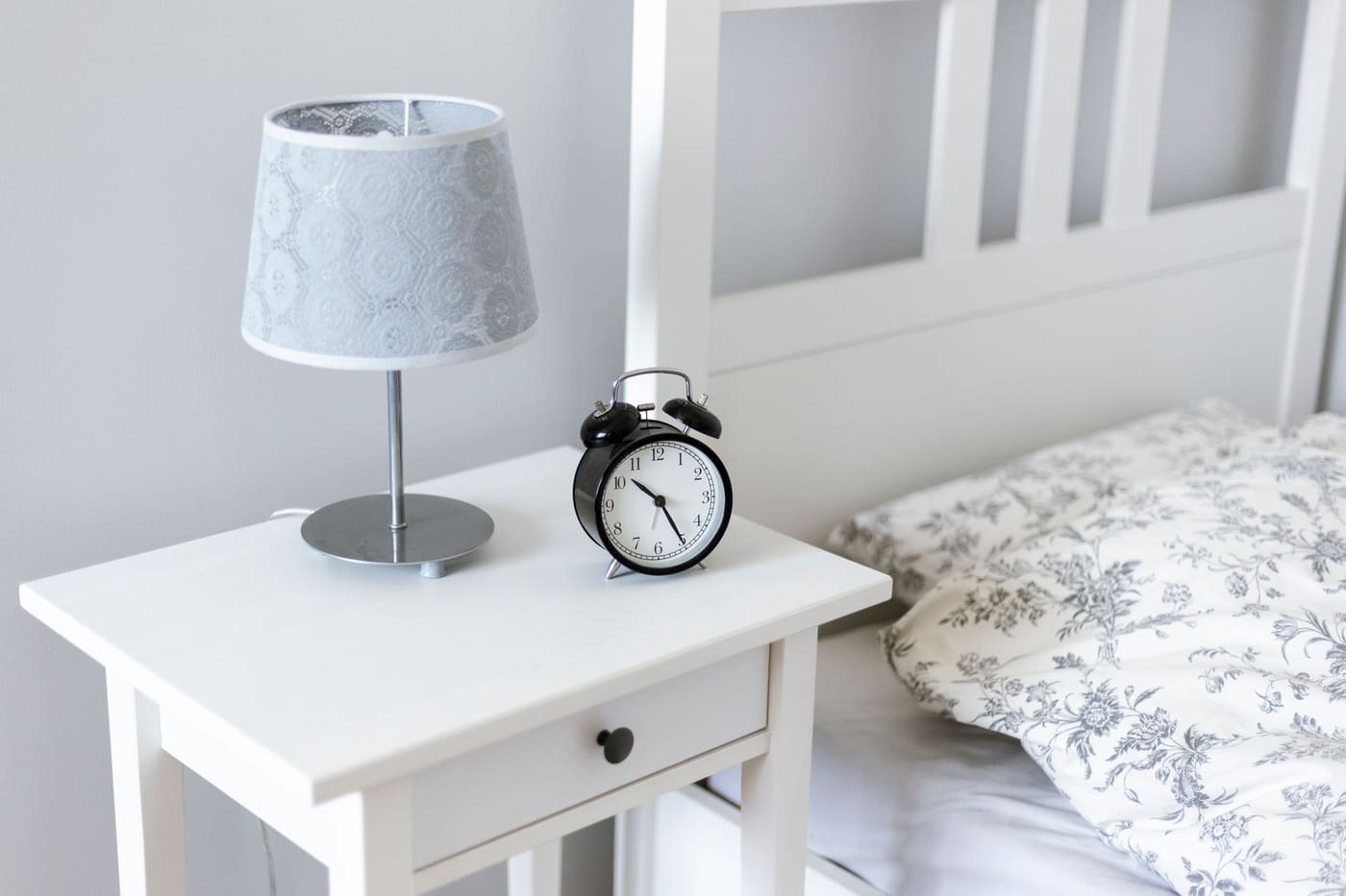 A white bedside table with a lamp and a small analog alarm clock beside a made bed