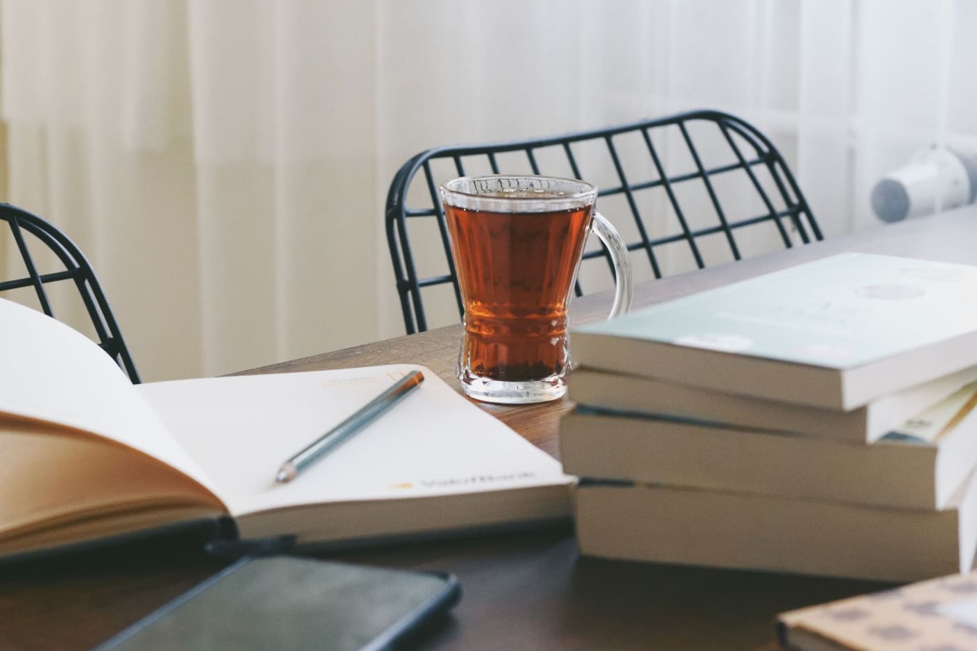 A glass of tea beside an open book and a stack of books on a table near a bright window