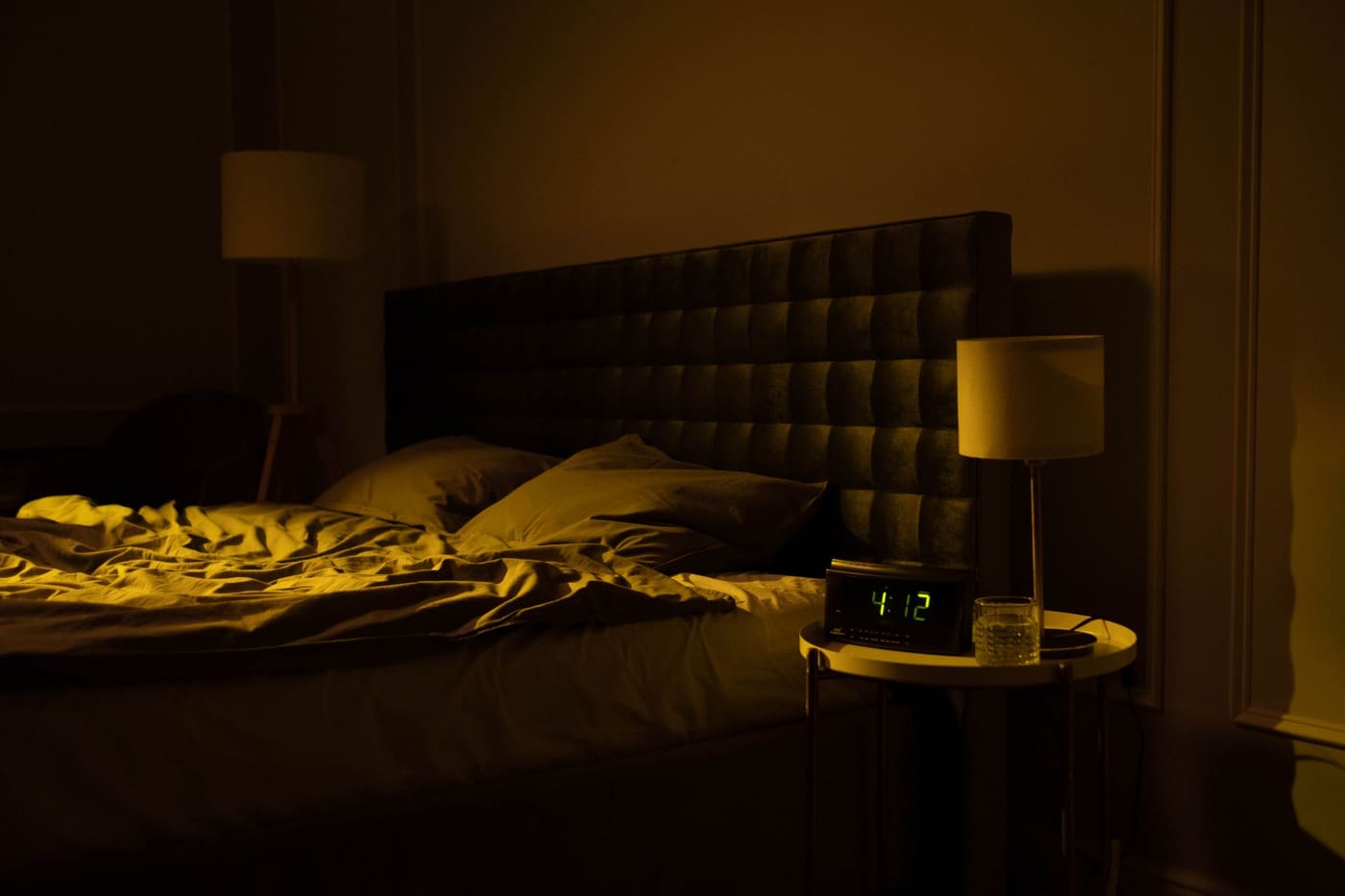 A dimly lit bedroom at night with an unmade bed, a lamp and a digital clock on the nightstand