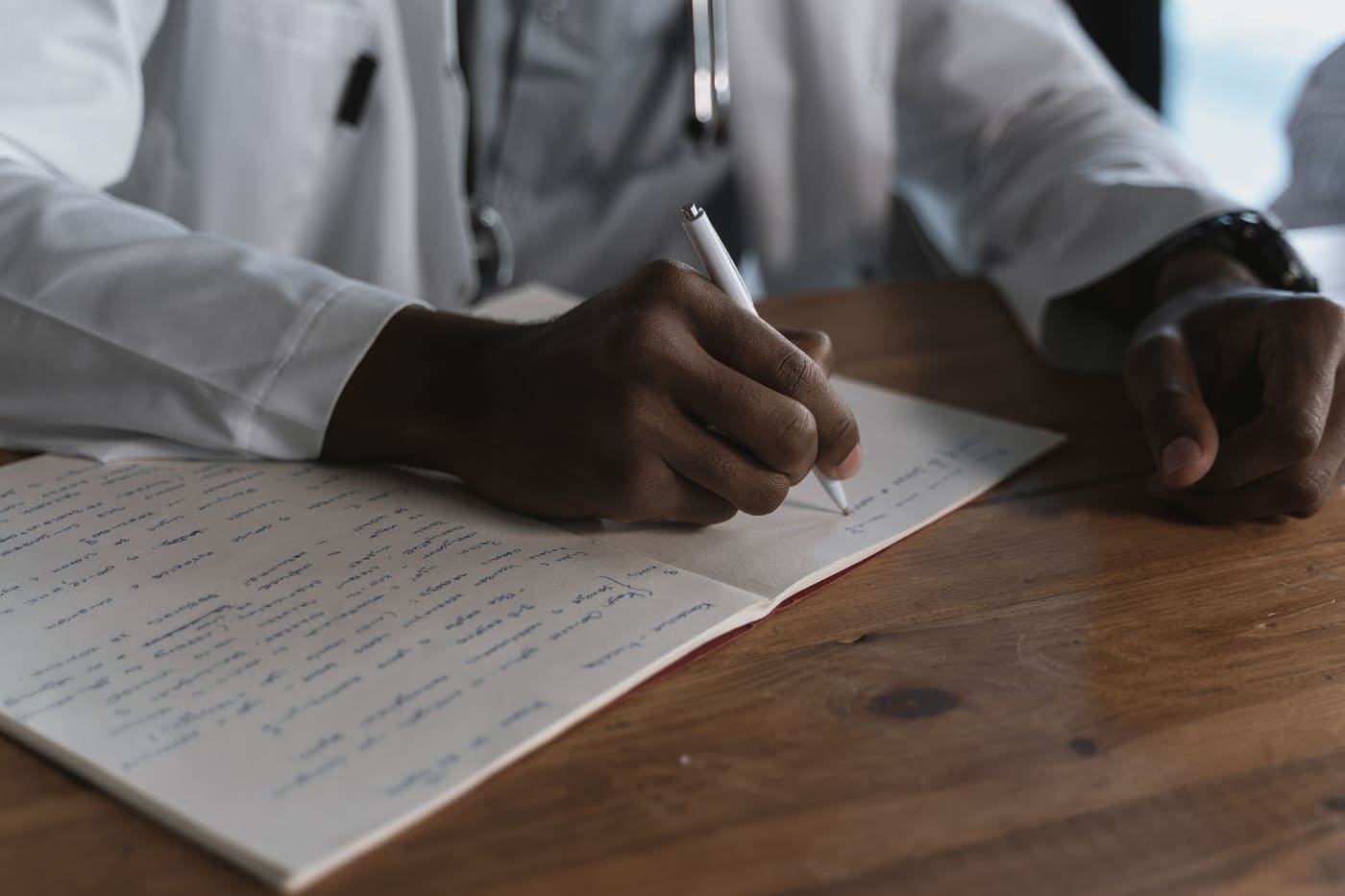 A doctor in a white coat writing notes in a notebook at a wooden desk