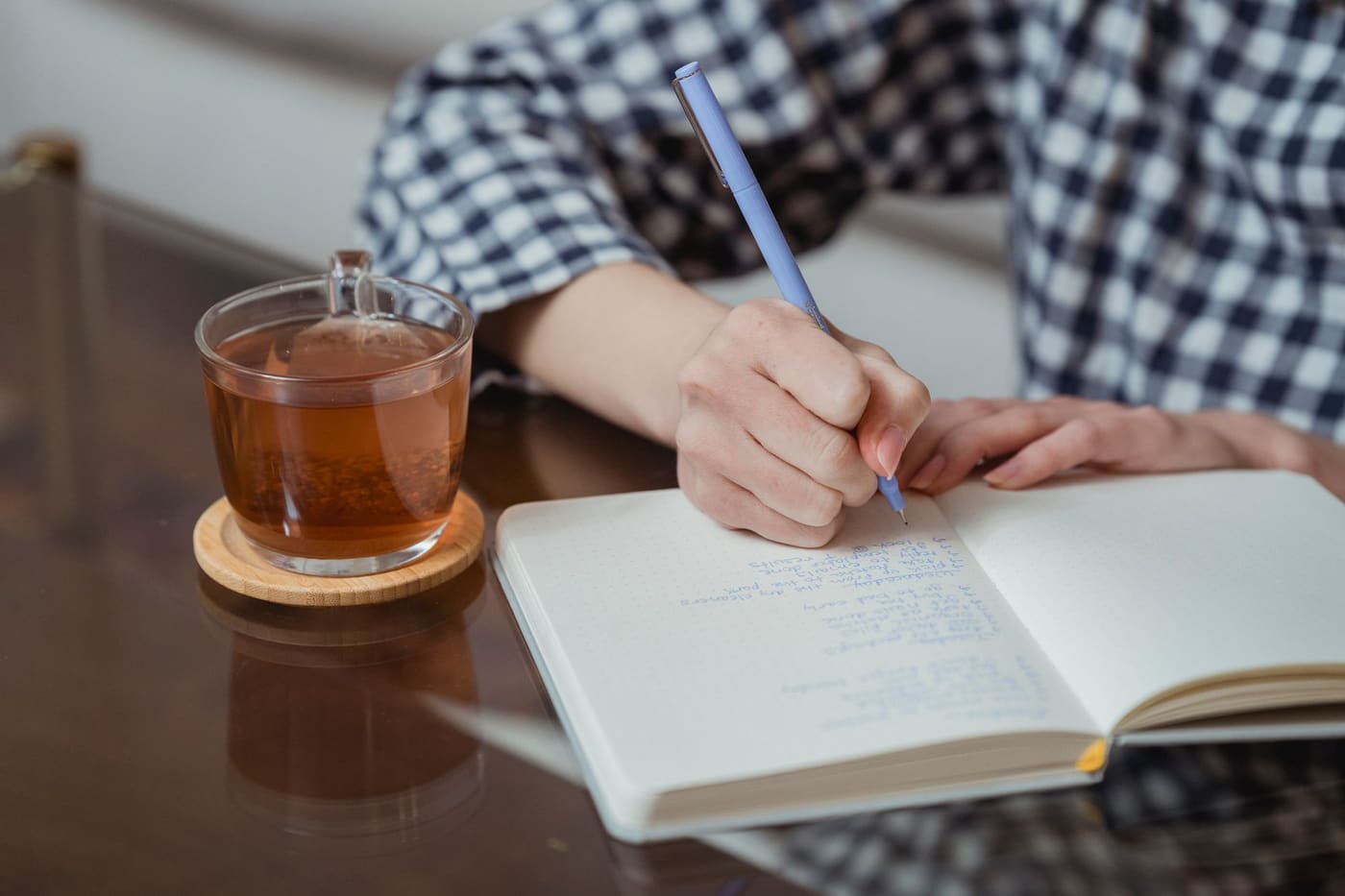 A person writing in a notebook at a table beside a glass cup of tea