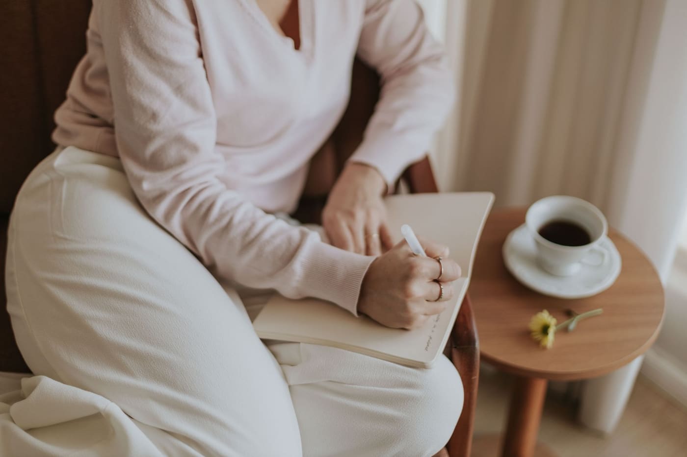 A person in a cream top writing in a notebook on their lap beside a small table with a cup