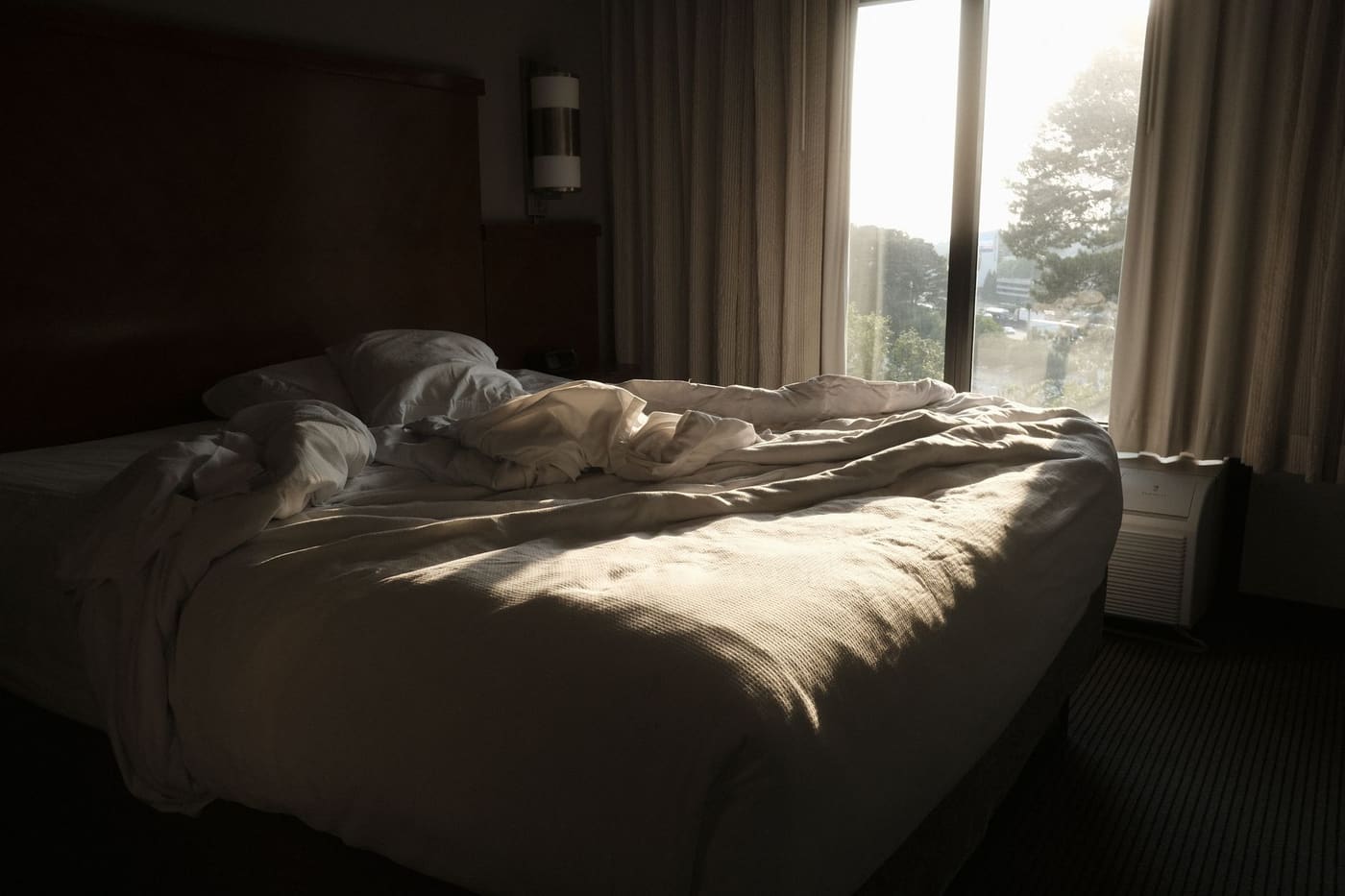 A bed with rumpled white sheets in a bedroom with morning sunlight coming through a large window