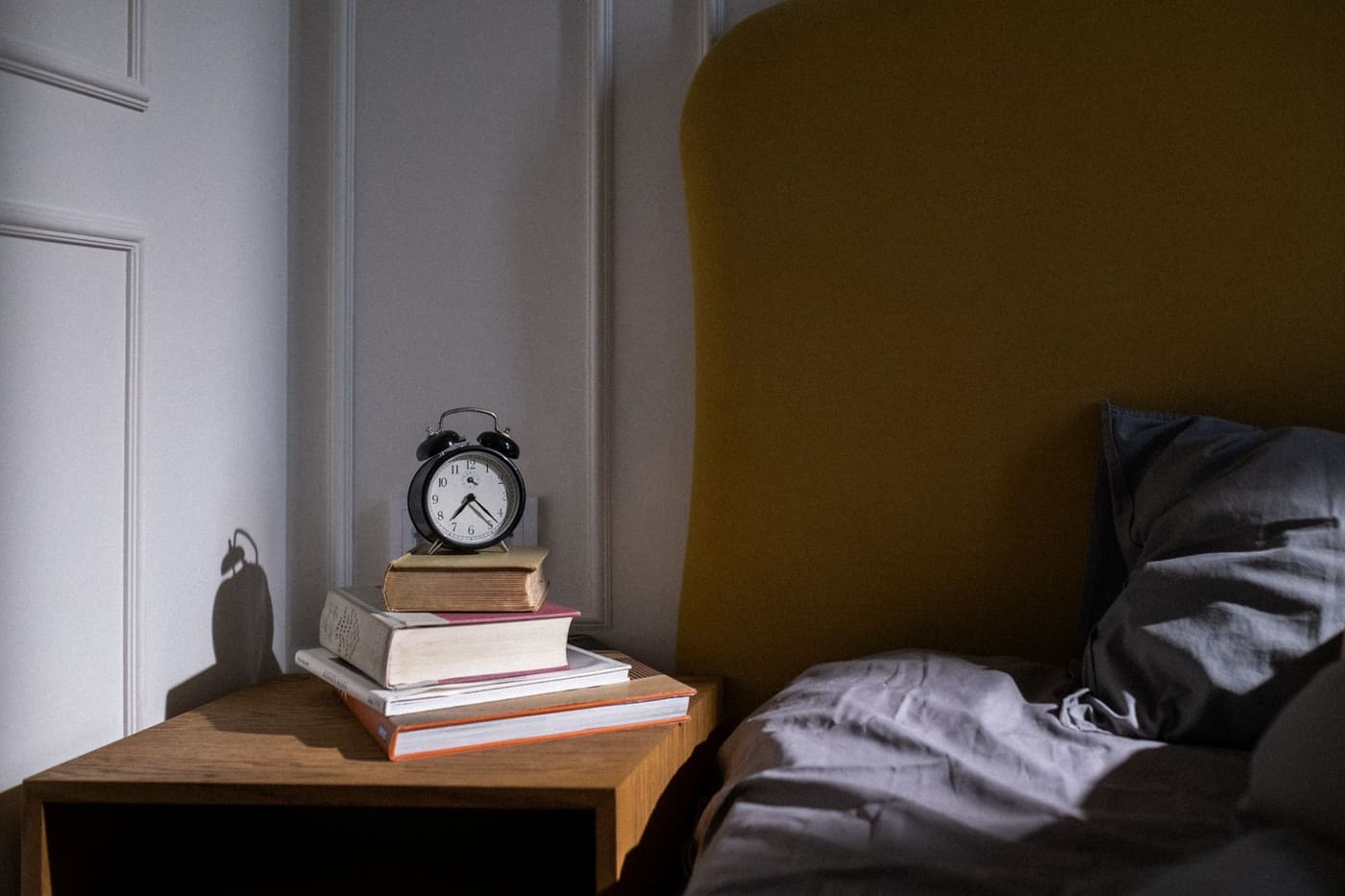 A wooden nightstand with a stack of books and a black alarm clock beside an unmade bed