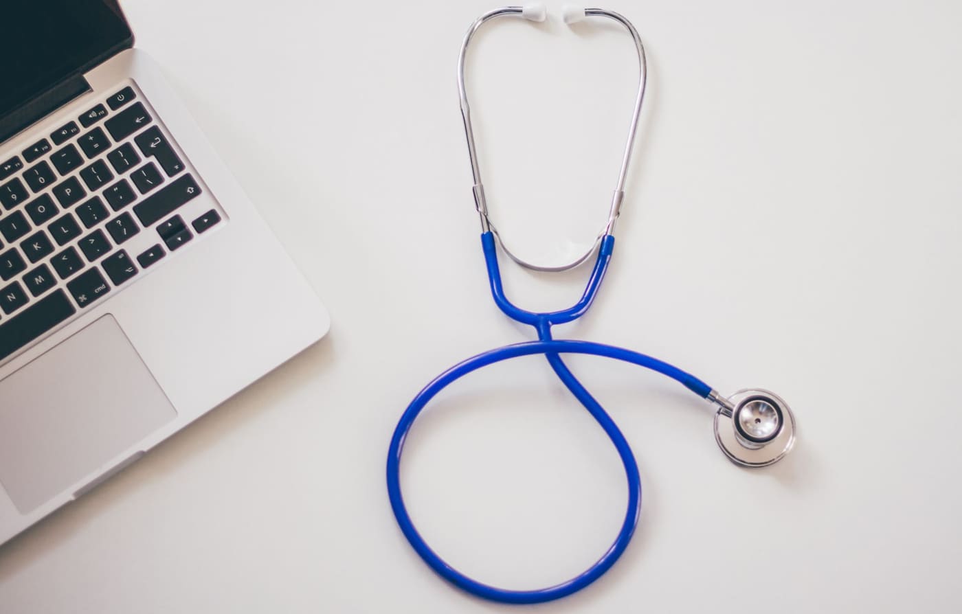 A blue stethoscope lying on a white desk beside a laptop